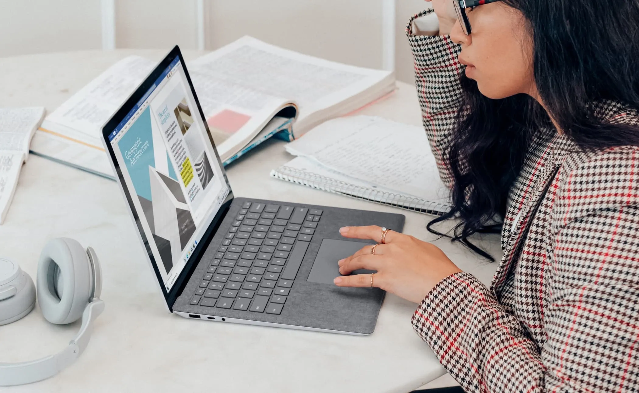 Woman working on a layout on her laptop, surrounded by open books, notes and headphones.