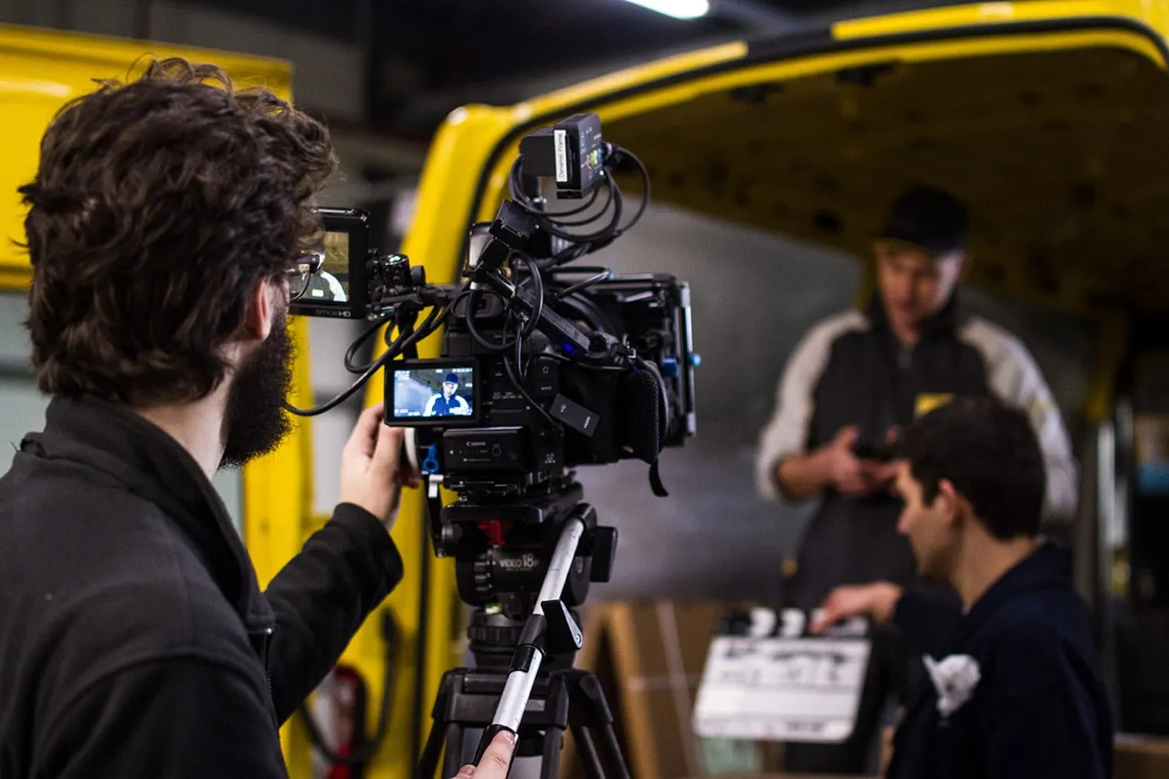Camera operator films two men in front of a yellow delivery van, a clapperboard held in the foreground.