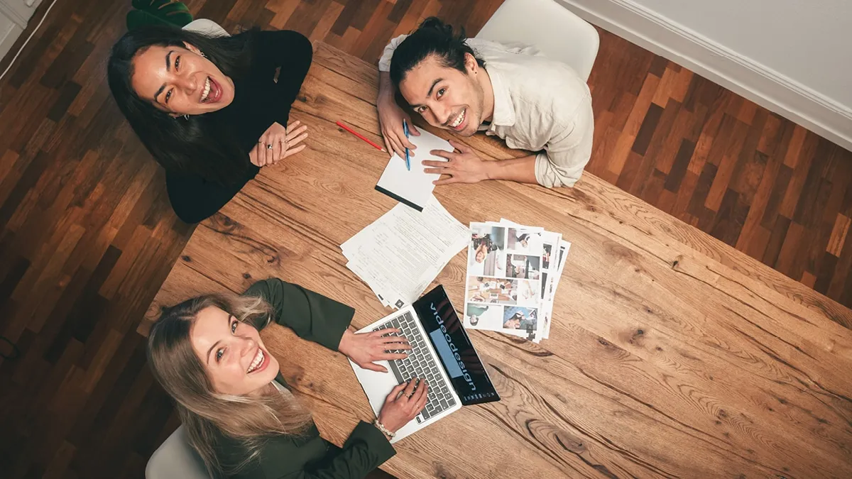 Overhead view of three people laughing around a wooden table with a laptop, storyboards and notes