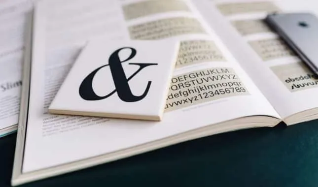 Open type specimen book showing alphabets, with a card bearing a large ampersand resting on it.