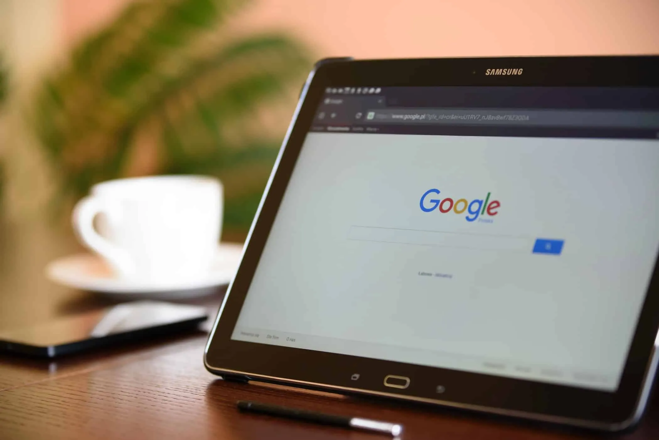 Tablet on a wooden desk with the Google search page open, next to a coffee cup and a stylus.
