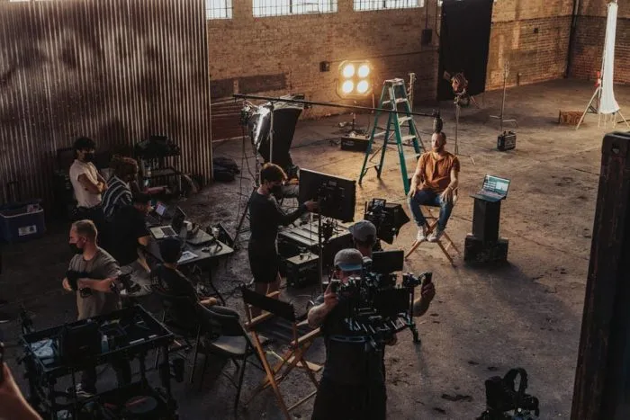 Filmcrew in einer Lagerhalle: Kameras, Scheinwerfer und Monitore rund um einen Darsteller auf dem Regiestuhl.