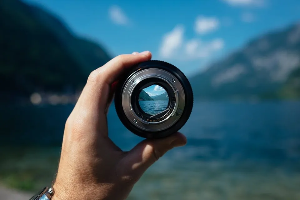 Hand holds a camera lens in front of a blurred mountain lake; the view through the glass is sharp.