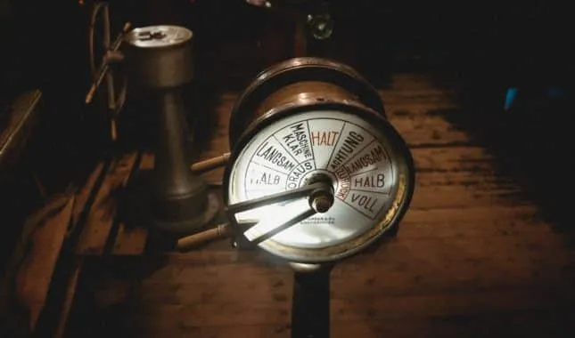 Brass engine-order telegraph on a ship's wooden deck, its dial marked with commands from stop to full ahead.