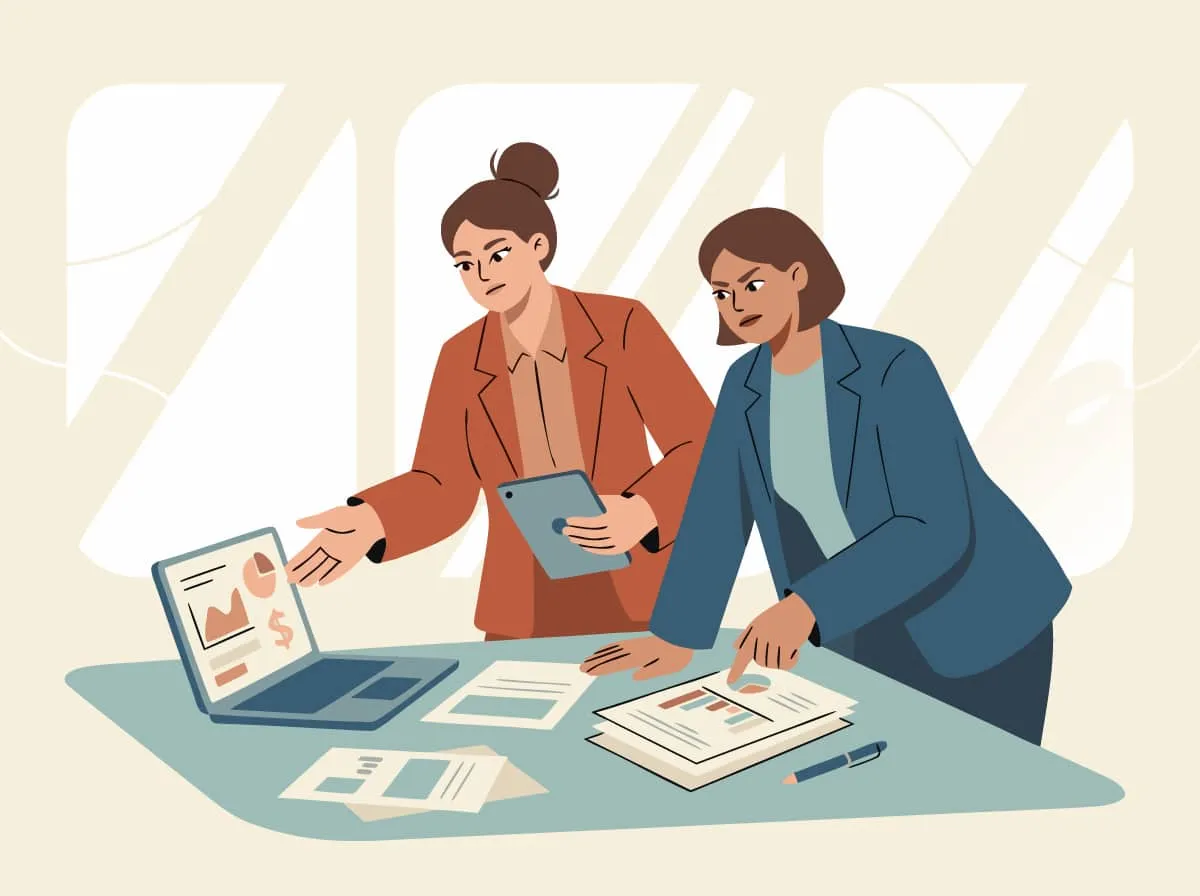 Two women lean over a laptop and printed reports, discussing the figures with sceptical faces.