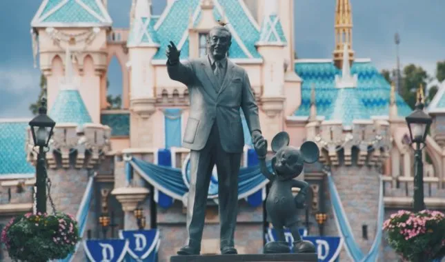 Bronze statue of Walt Disney holding Mickey Mouse's hand in front of the Disneyland fairy-tale castle.