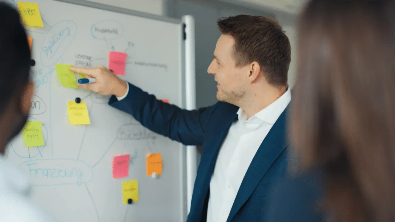 Man in a suit explains a whiteboard mind map covered in coloured sticky notes to colleagues in the foreground.