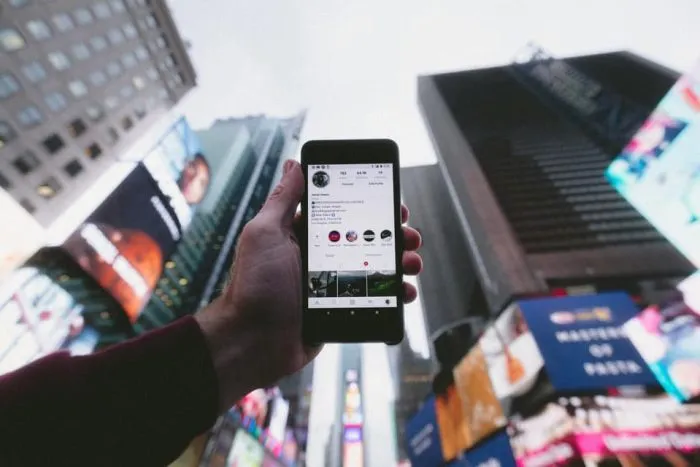Hand hält ein Smartphone mit Instagram-Profil vor Hochhäuser und riesige Werbebildschirme