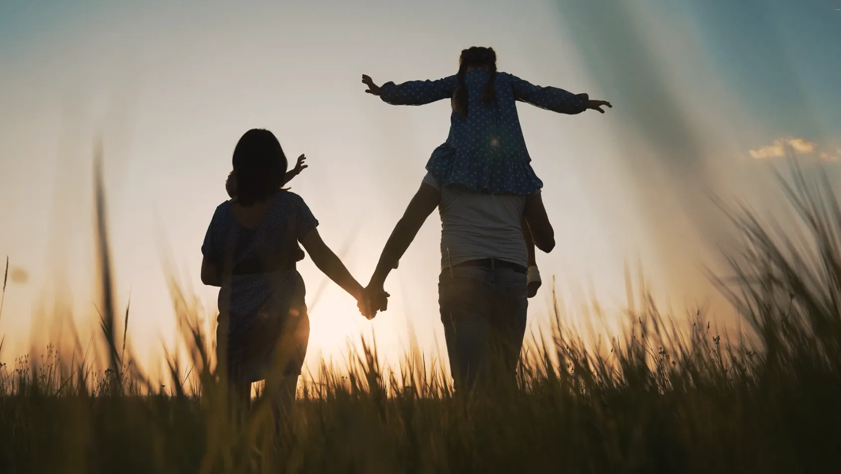 Backlit family walking through tall grass at sunset, the daughter riding on her father's shoulders