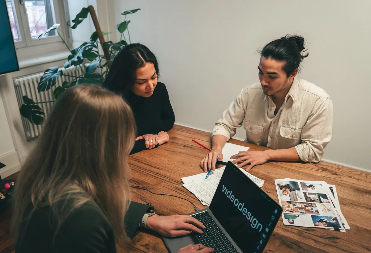 Three people discuss printed storyboards at a wooden table, with an open laptop and handwritten notes beside them.