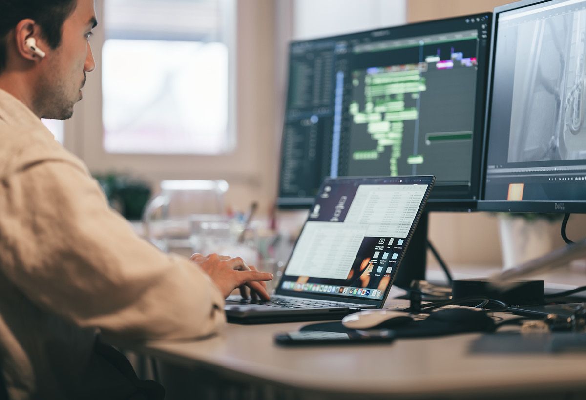 Colleague with an earbud working at a laptop and two monitors