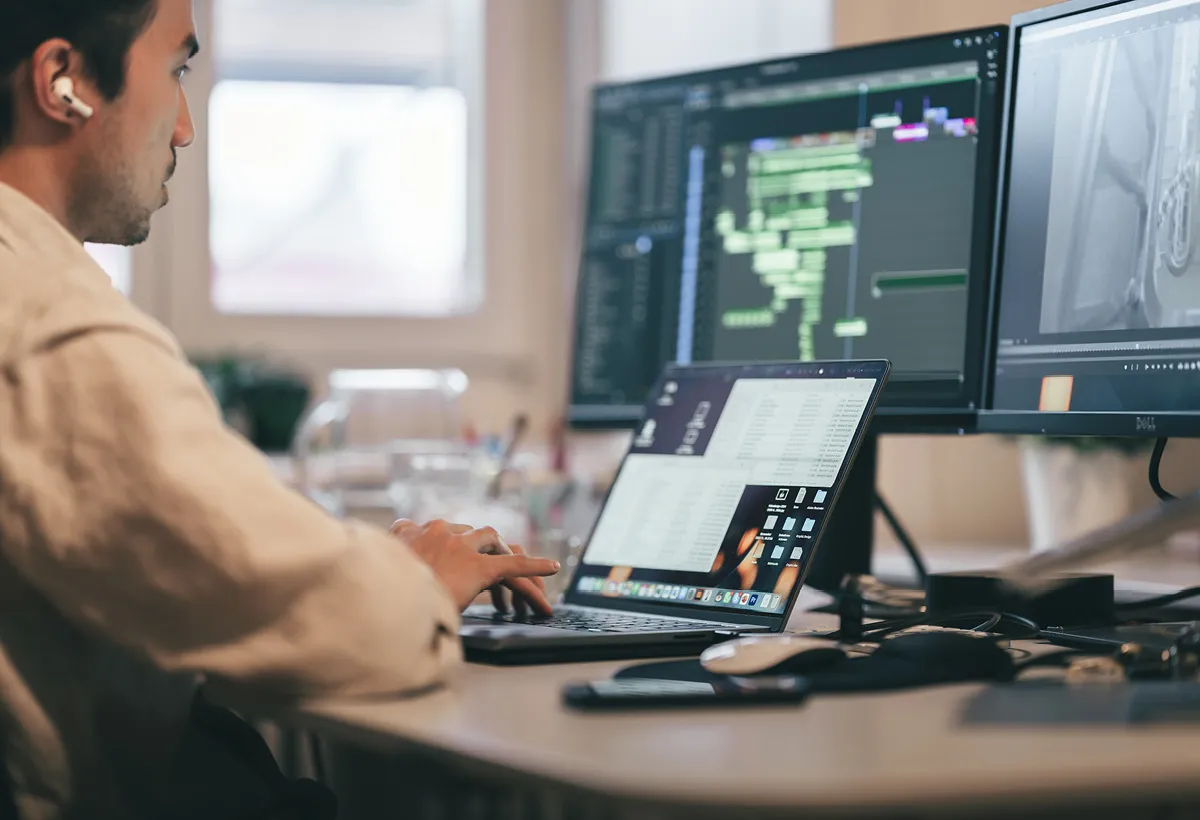 Editor at a desk works on a video: an editing timeline on the monitor and a list of figures on the laptop.